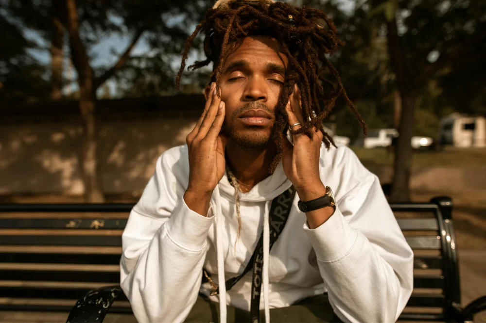 A man with locs in a knot on top of his head wears a white hoodie and sits on a bench. His eyes are closed and his fingers touch his cheeks and he leans into his hands.