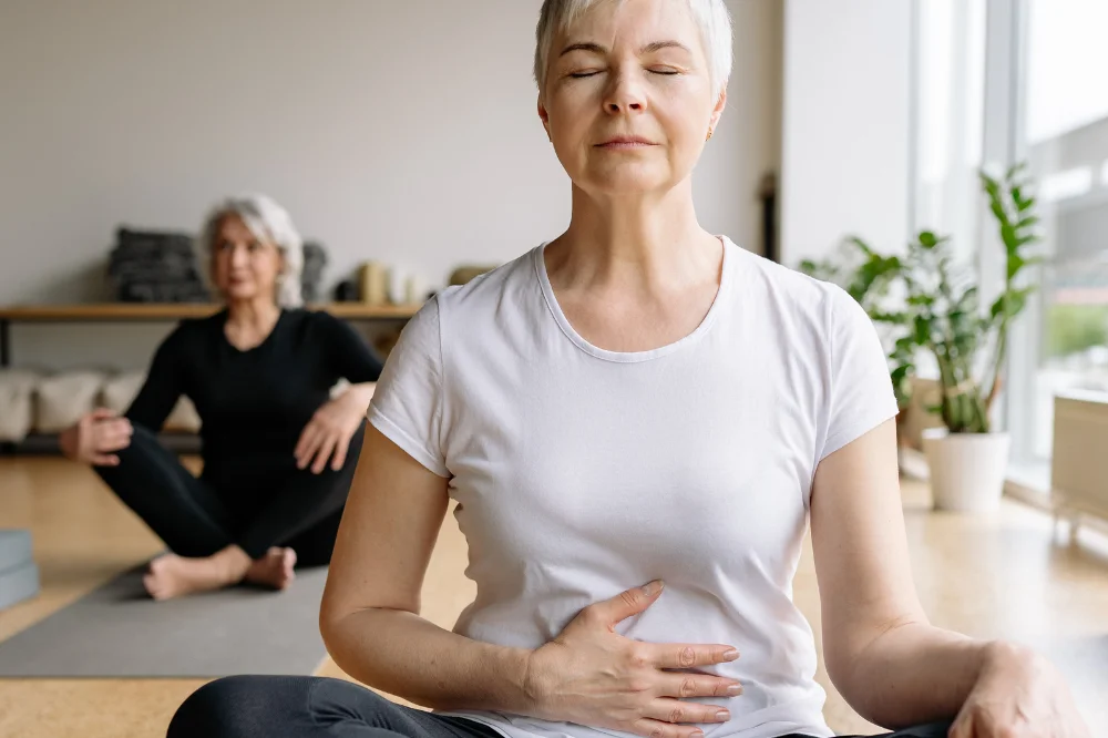 a woman with short white hair and a white shirt sits cross legged on a yoga mat . Her eyes are closed and her hand is on her stomach.