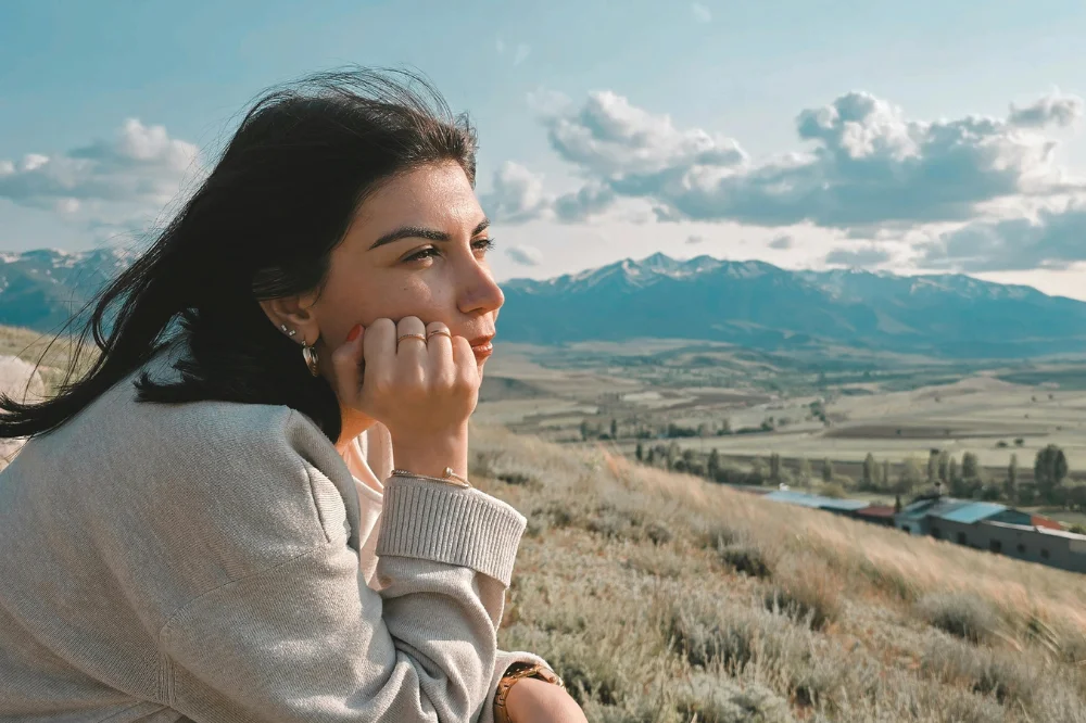 A side view woman with shoulder length dark hair leans her chin into her hand and looks into the distance. There are mountains behind her.