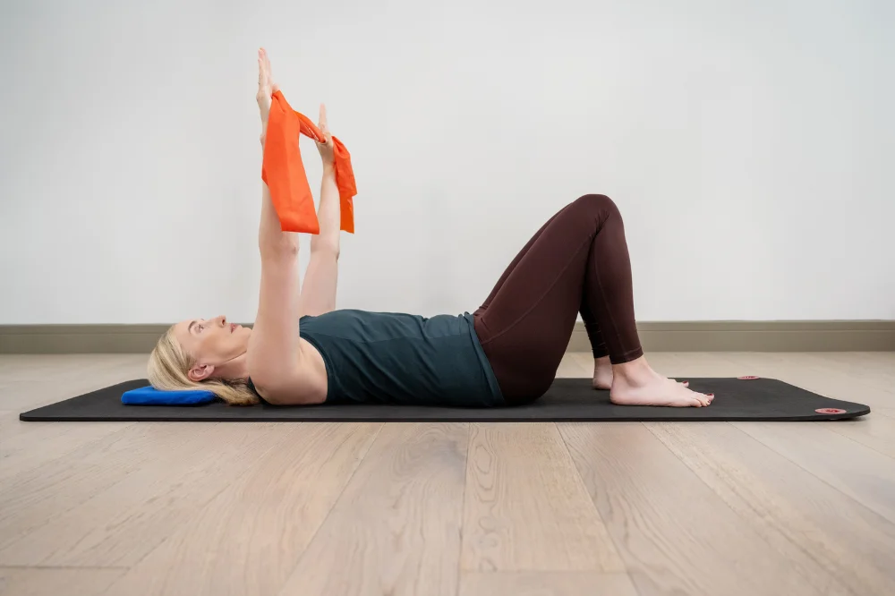 Jeannie demonstrates a shoulder stability exercise. She is on her back on her mat with her knees up, she has a theraband between both hands that are extended into the air above her.