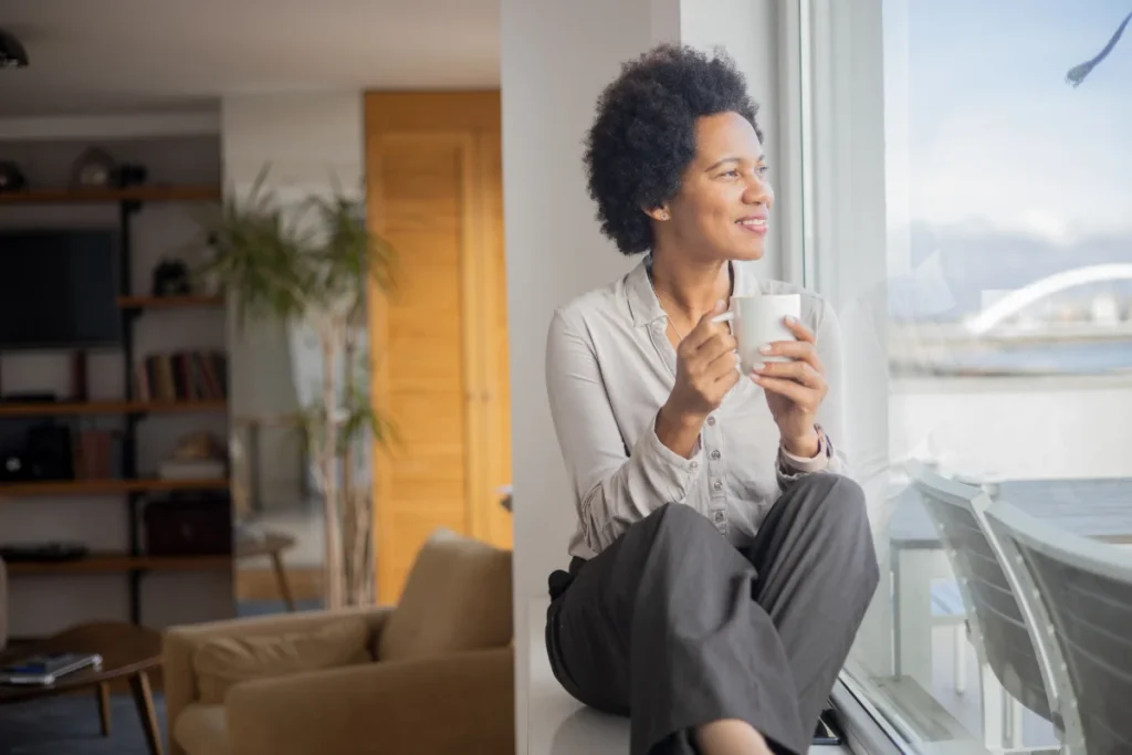 a woman holds a coffee cup and sits on a windowsill looking out a large window