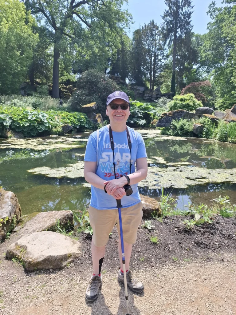 Ian stands in front of a scenic pond with rocks and trees in the background. He holds a walking stick and has his camera on his neck. 