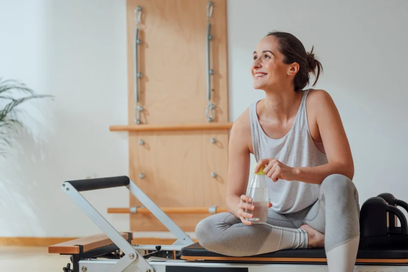 a woman sits on a reformer with one leg under her looking off to the side smiling and drinking water 