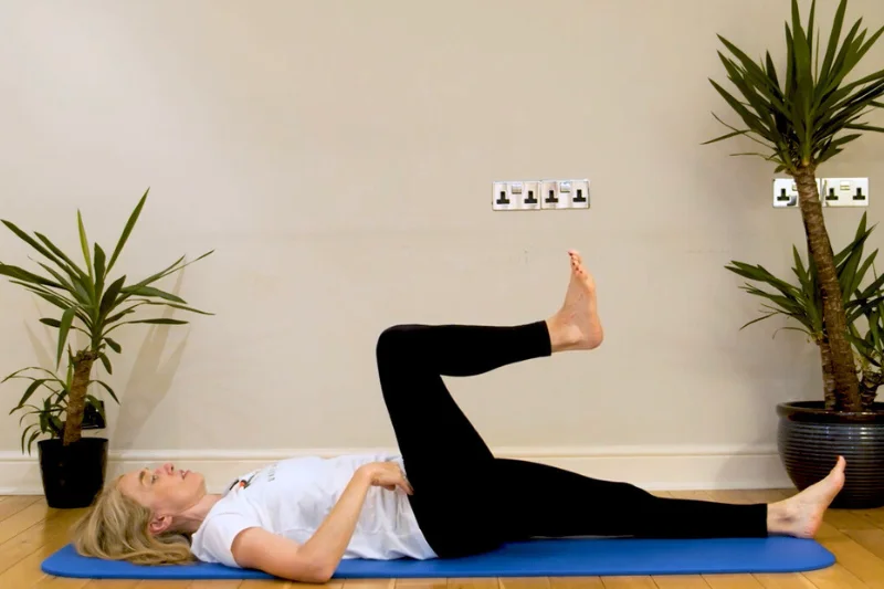 Jeannie di Bon laying supine on a yoga mat with one bent leg in the air 
