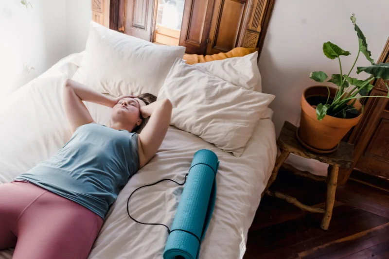 woman in exercise clothes looks tired laying on the bed with her yoga mat rolled up next to her, her hands are on her head 