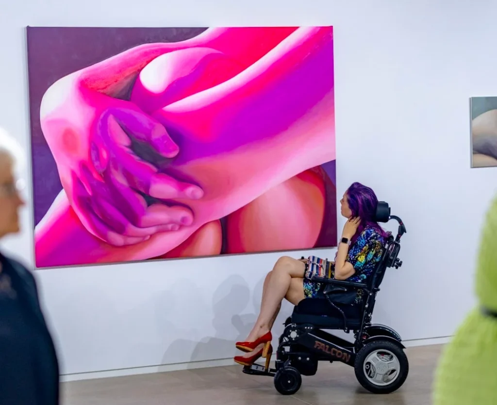 Christy sits in a wheelchair with her legs crossed and red high heels looking at a painting of a person's arms crossed over their legs, painted in pinks and purples.