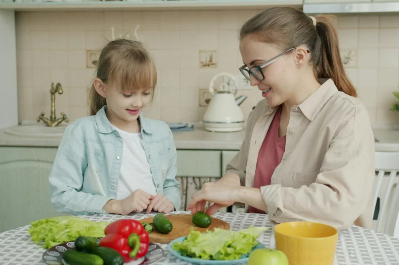 A woman sits at a table chopping vegetables, a little girl stands next to her watching both are smiling