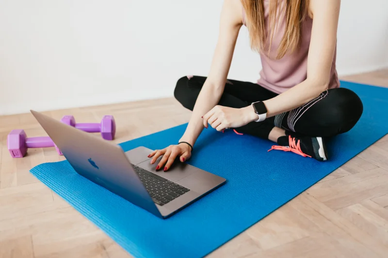 woman sits crosslegged on a yoga mat with her fingers on the mouse pad of a laptop preparing to do an exercise video