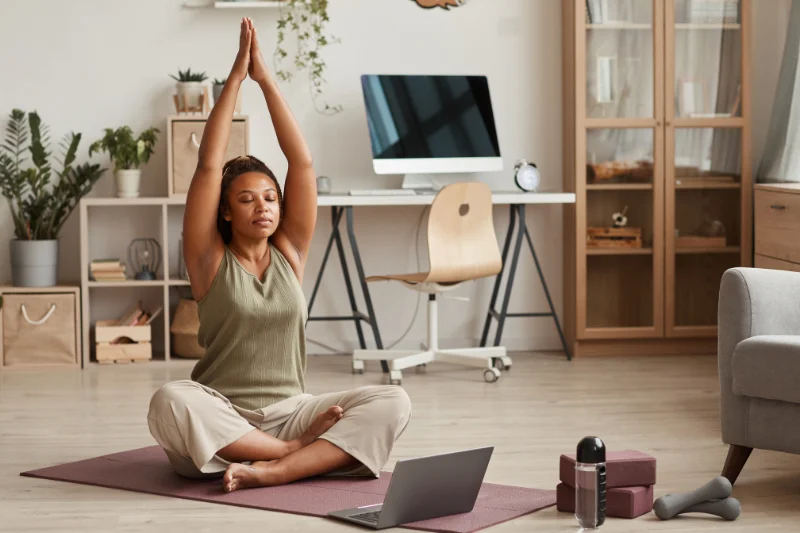 a woman sits crosslegged on her yoga mat with her hands joined stretching over her head with a laptop in front of her like she is doing an exercise video