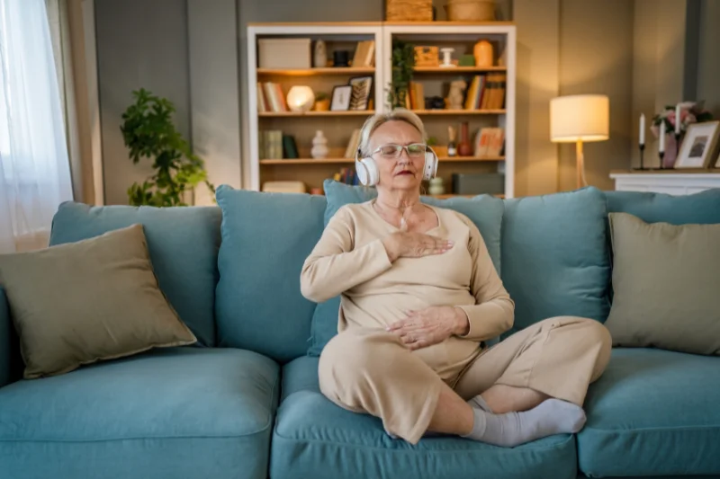 An older woman sits on the couch with her legs crossed. She is wearing headphones with her eyes closed, with one hand on her chest and one on her stomach, appearing ot meditate