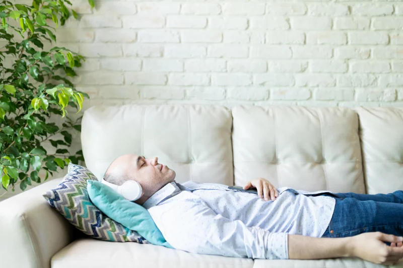 A man is laying on a couch with his head supported. He is wearing headphones and his eyes are closed as he meditates.