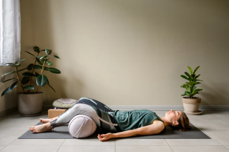 A woman lays on her back on a yoga mat with her arms by her side and her knees resting over a bolster in relaxation
