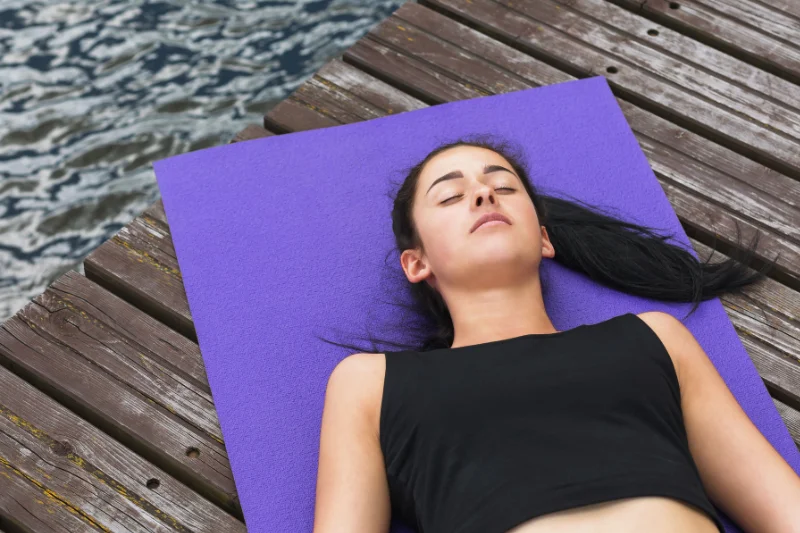a woman lies on a yoga mat on her back with her eyes closed. She is lying on a wooden dock with water visible beyond it.