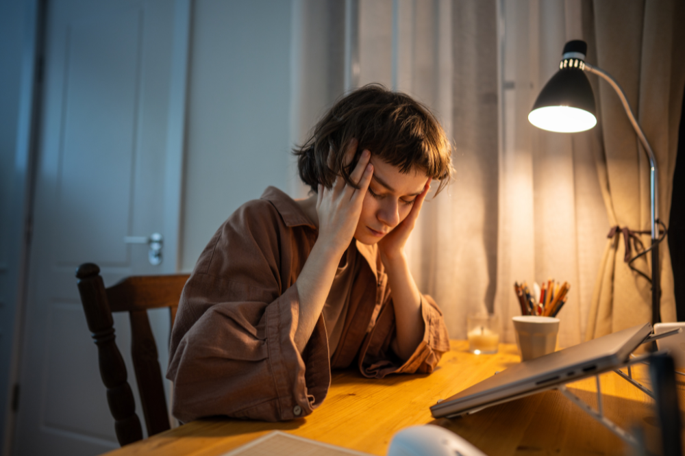 A woman sits at a desk with her eyes closed and hands on the side of her head looking fatigued