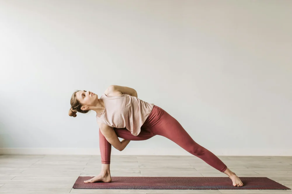 A woman on a yoga mat is doing a pose where she connect her arm behind her under one leg.
