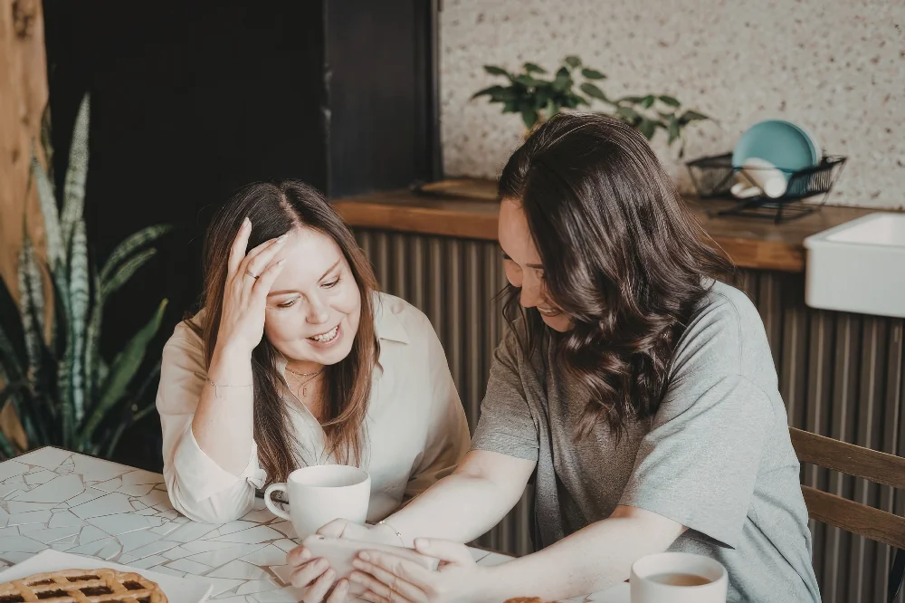 Two woman sit at a table together laughing and looking at a phone experiencing community together