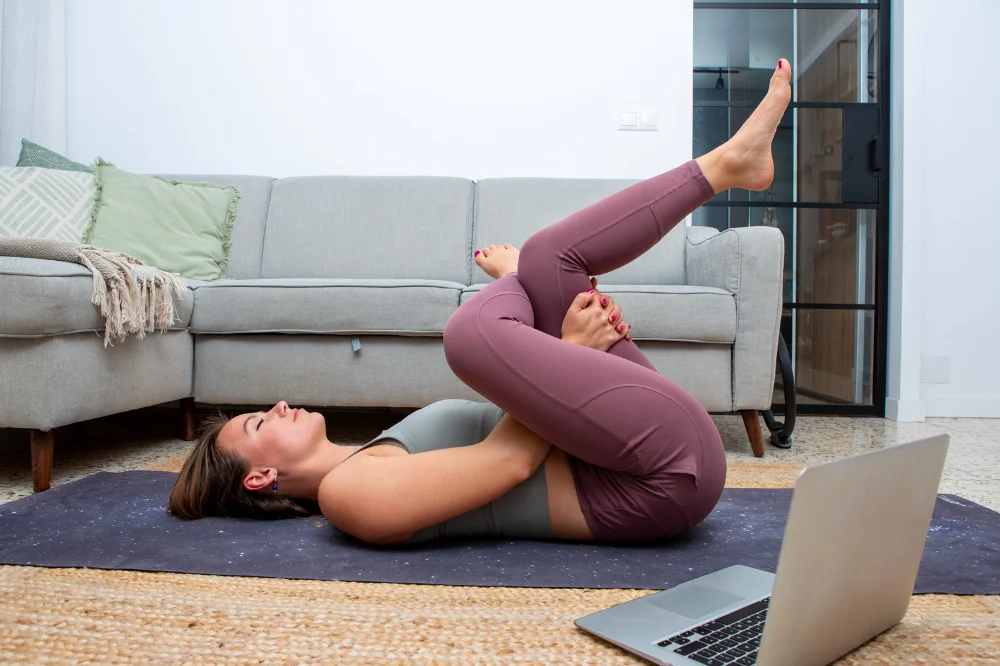 A woman lays on a yoga mat in her living room doing a figure four stretch, with a laptop open beside her.